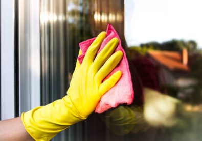 A hand in yellow protective glove cleaning the glass window pane with a pink rag. Home routine.